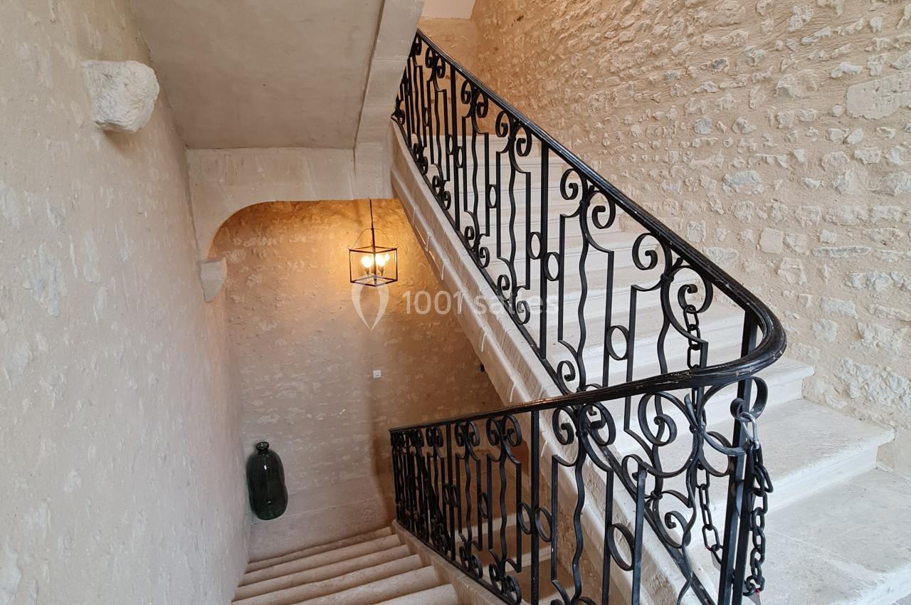 Escalier en pierre avec rampe en fer forgé, éclairé par une suspension, dans un intérieur aux murs en pierre apparente.