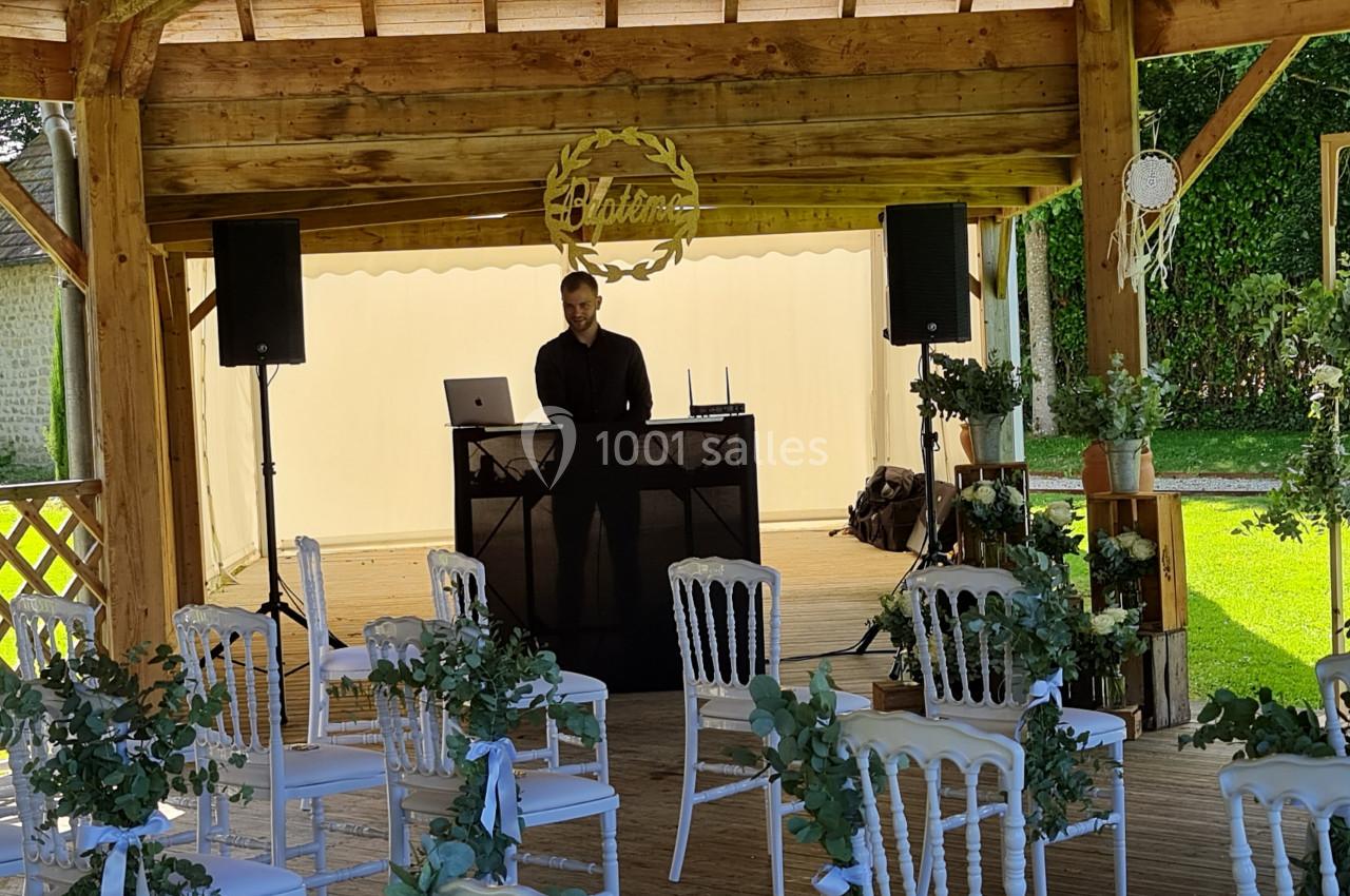 Un DJ se tient derrière sa table sous une pergola en bois décorée pour un événement avec des chaises blanches et des fleurs.