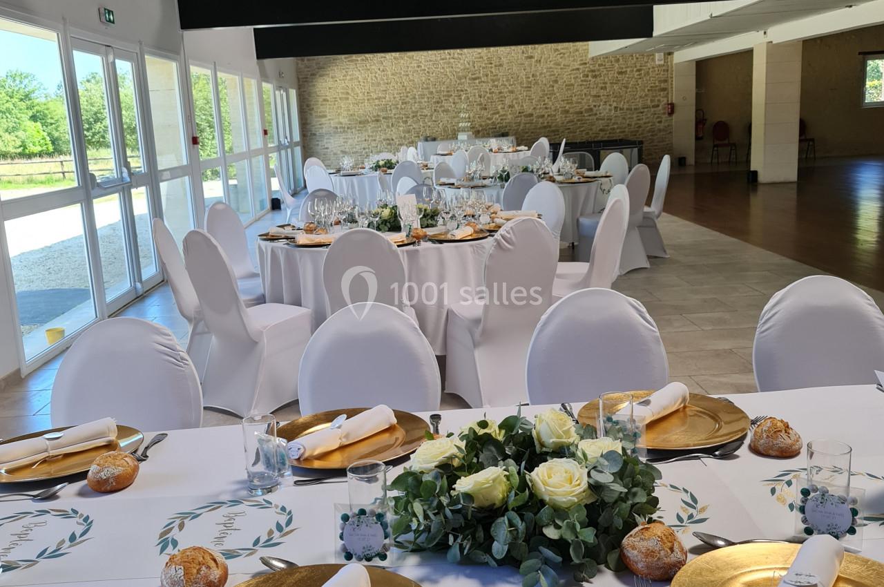 Salle de réception lumineuse avec tables rondes dressées, nappes blanches, assiettes dorées et décoration florale.