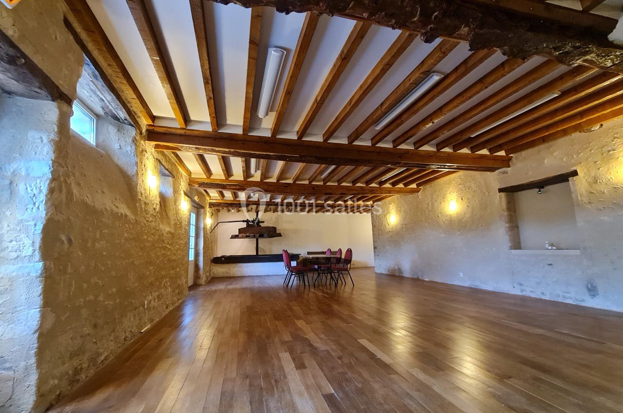 Salle spacieuse avec murs en pierre, poutres apparentes, parquet en bois et une table entourée de chaises au centre.