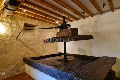 Pressoir ancien en bois dans une pièce avec murs en pierre, plafond à poutres apparentes et sol en parquet.