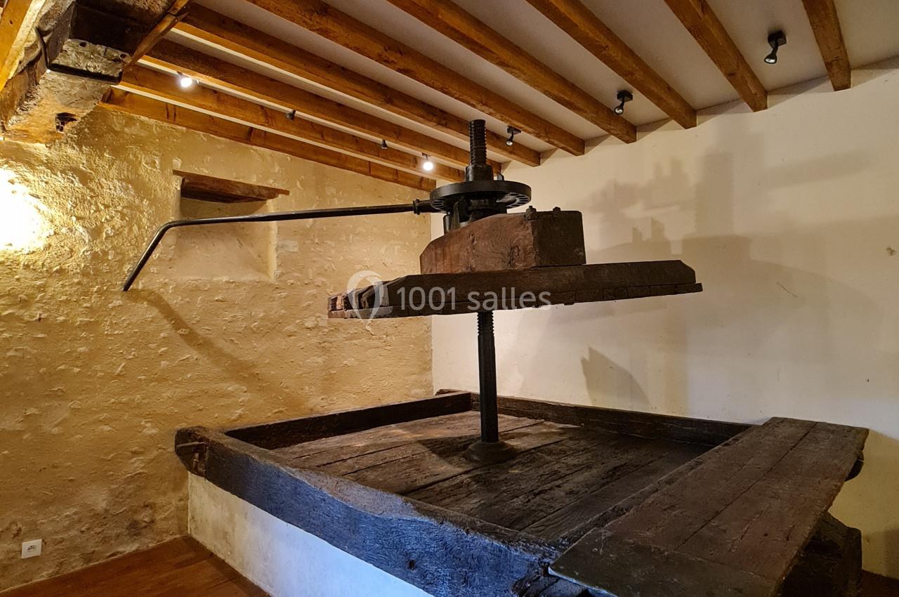 Pressoir ancien en bois dans une pièce avec murs en pierre, plafond à poutres apparentes et sol en parquet.