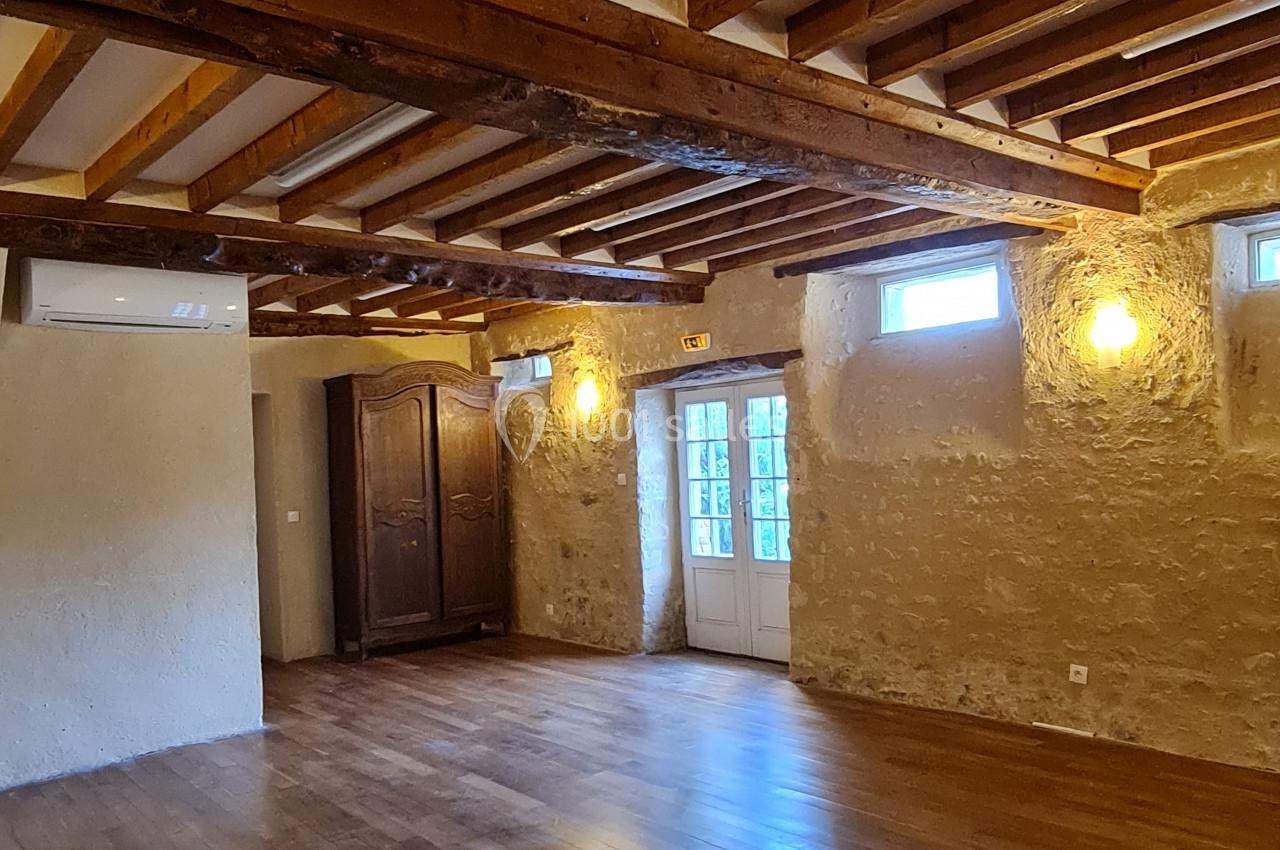 Salle avec parquet en bois, murs en pierre apparente, poutres au plafond, éclairage mural et porte-fenêtre vitrée.