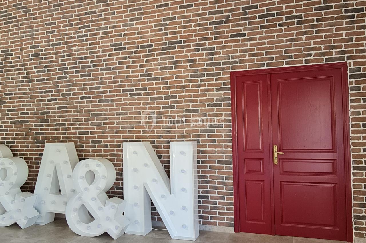 Mur de briques avec une porte rouge à double battant et des lettres lumineuses blanches posées au sol.