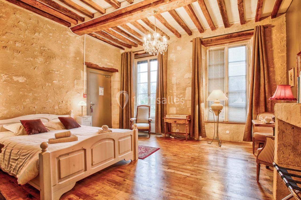 Chambre lumineuse avec lit double, poutres apparentes, murs en pierre, parquet et mobilier classique.
