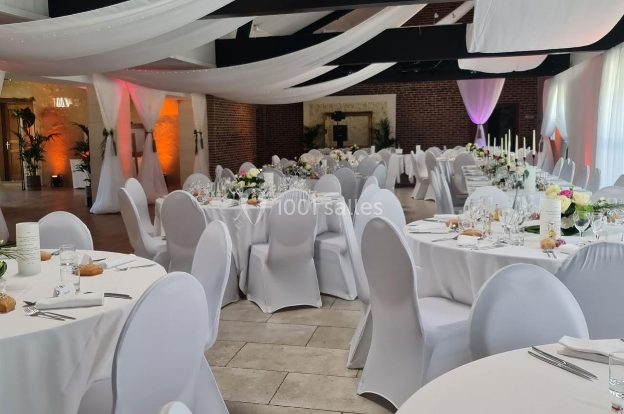 Salle de réception décorée avec des tables rondes dressées, nappes blanches et voilages suspendus au plafond.
