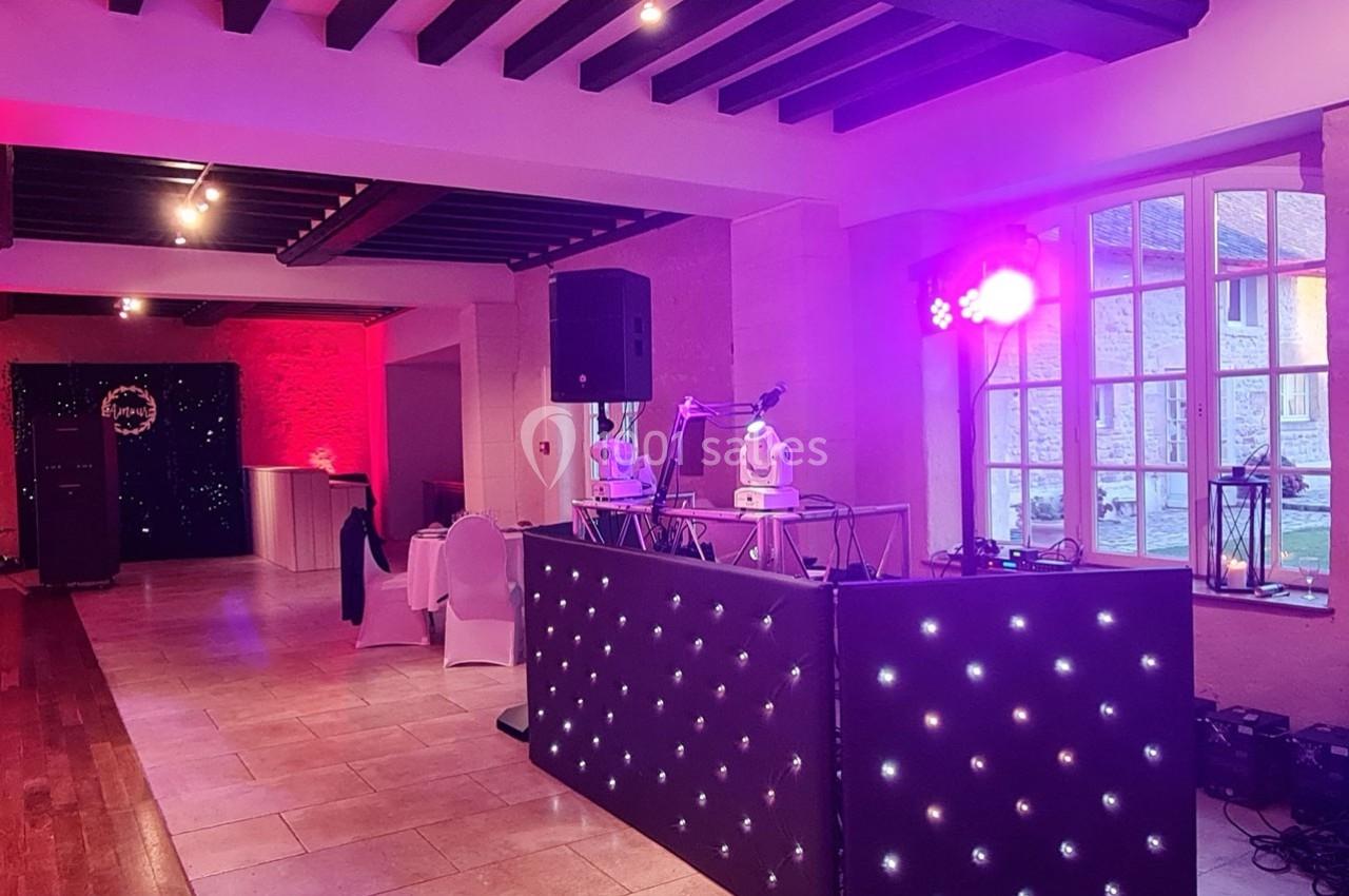 Salle de réception avec éclairage coloré, table de DJ équipée et chaises disposées près d'une grande fenêtre.