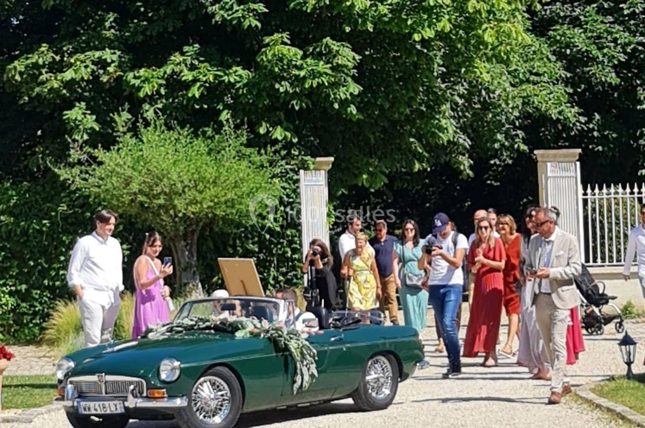 Une voiture décapotable verte décorée de fleurs arrive devant un groupe de personnes élégamment vêtues en extérieur.