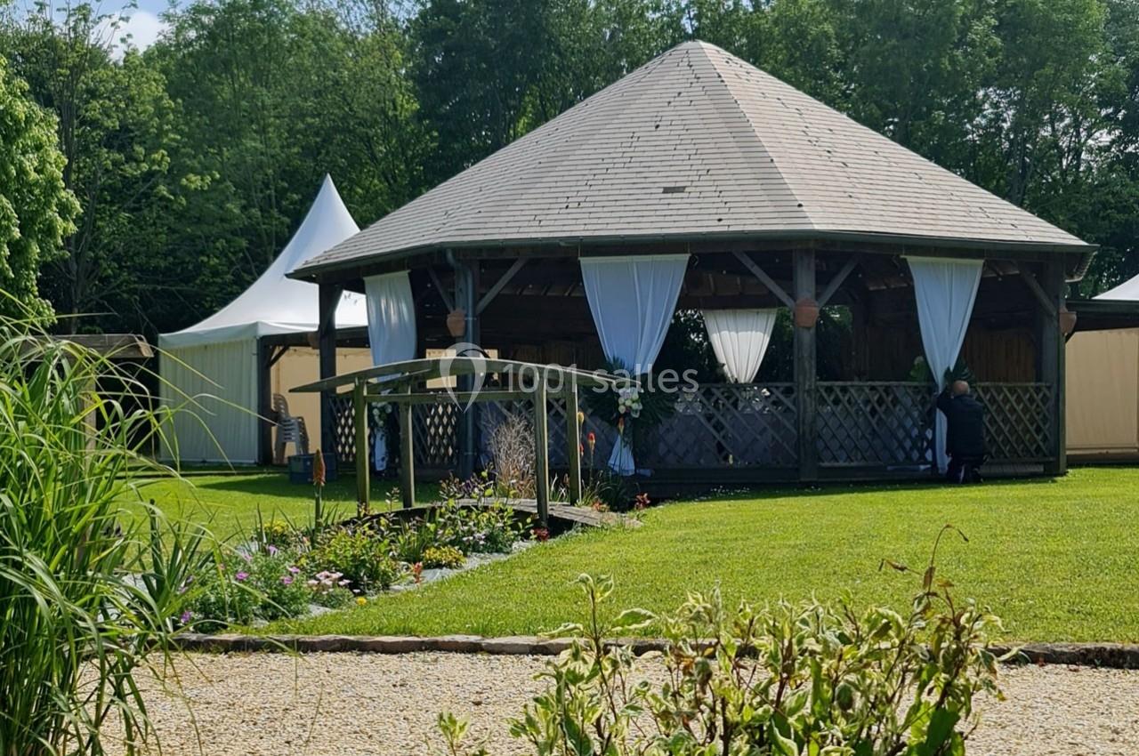 Pavillon en bois avec rideaux blancs entouré de pelouse, fleurs et arbres, sous un ciel dégagé.