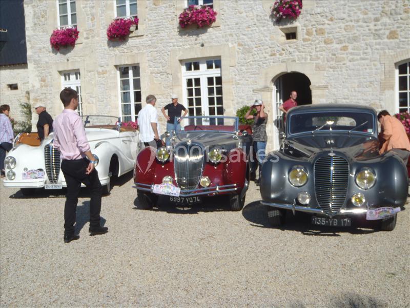 Trois voitures anciennes exposées devant un bâtiment en pierre, avec des personnes discutant autour.