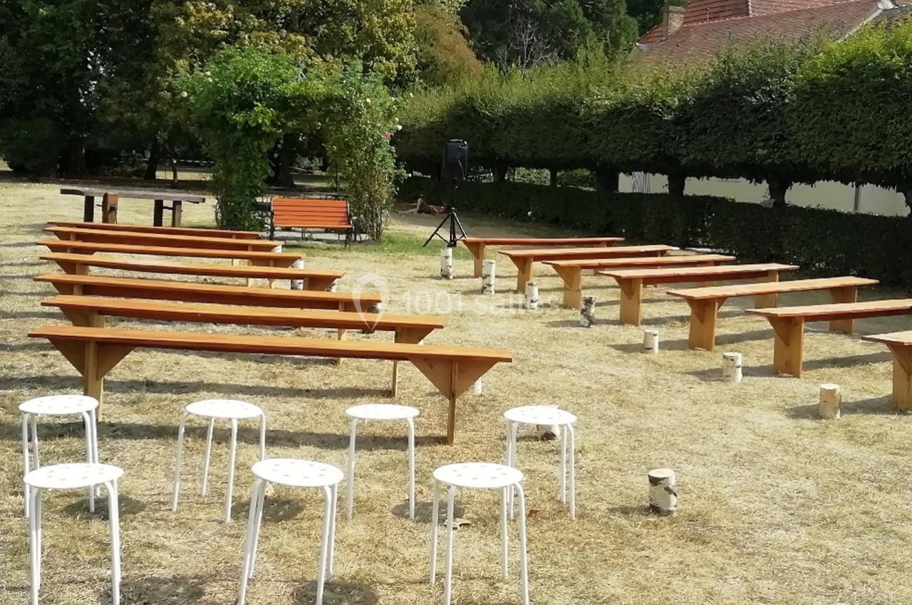 Bancs en bois et tabourets blancs disposés en extérieur sur une pelouse sèche, entourés de haies et d'arbres.