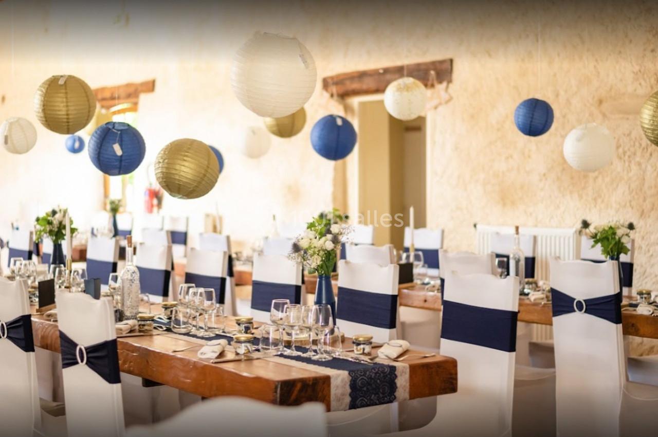 Salle de réception décorée avec des tables en bois, chaises blanches ornées de rubans bleus et lanternes suspendues.