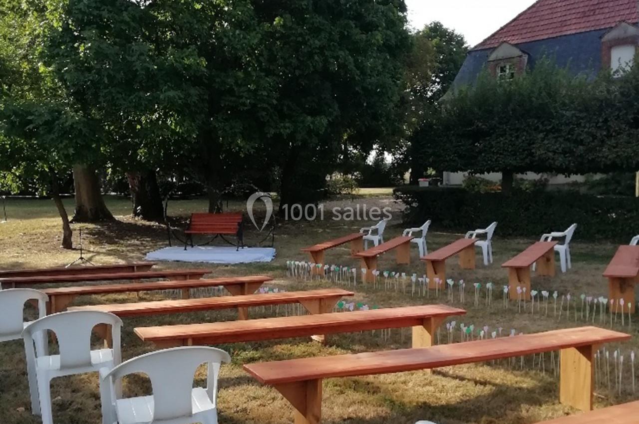 Bancs en bois et chaises blanches disposés en extérieur, devant un espace dégagé avec des arbres et une maison en arrière…