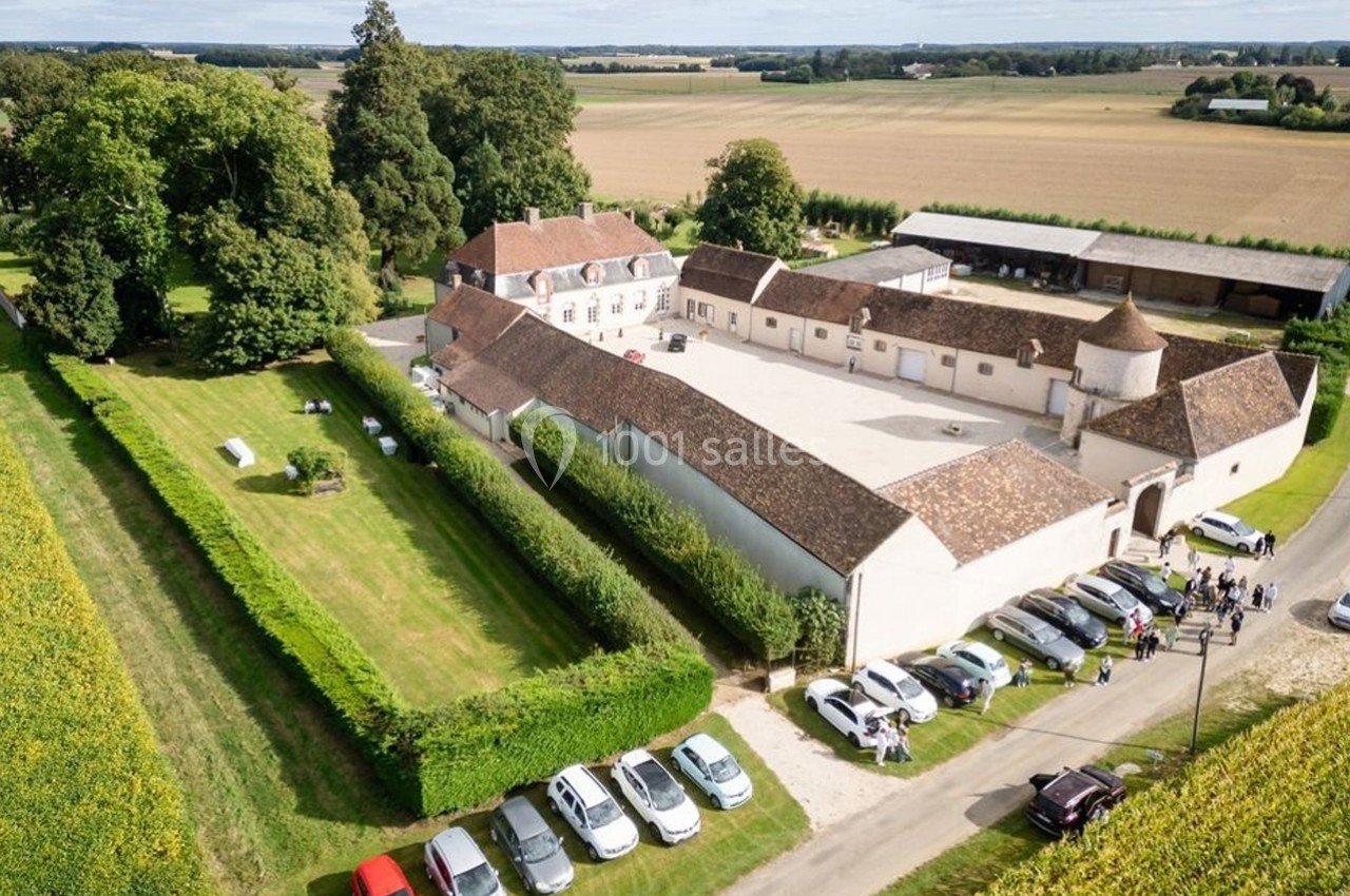 Vue aérienne d'un domaine rural avec bâtiments en pierre, cour centrale, champs environnants et voitures stationnées.