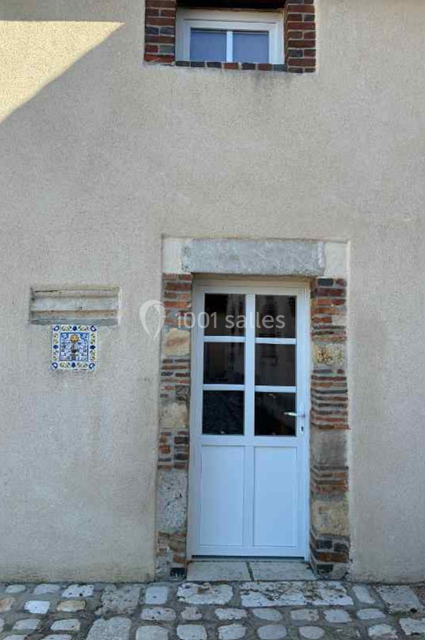 Porte blanche vitrée encadrée de briques et de pierres, avec une plaque décorative en céramique sur un mur beige.