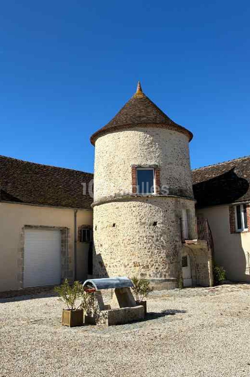 Bâtiment en pierre avec une tour ronde et un toit conique, entouré d'une cour en gravier sous un ciel bleu.