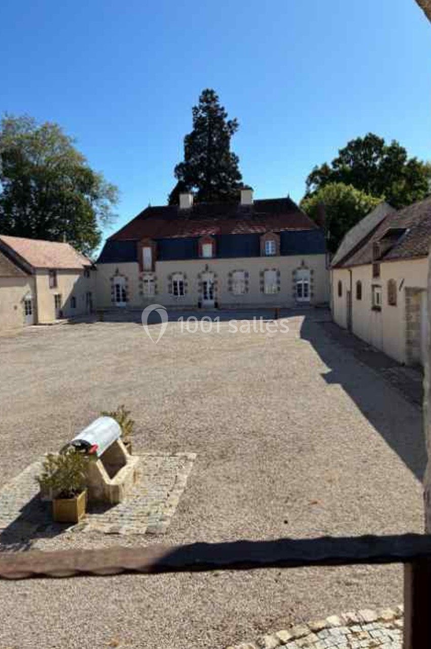 Cour pavée avec un bâtiment principal ancien entouré de dépendances, sous un ciel bleu dégagé.