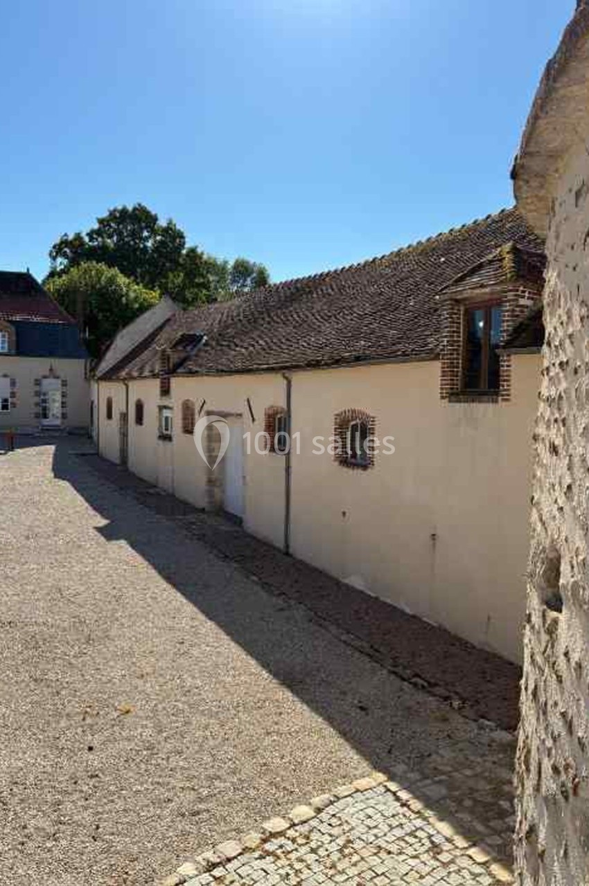 Bâtiments anciens en pierre et crépi avec toits en tuiles, sous un ciel bleu, dans une cour pavée et gravillonnée.