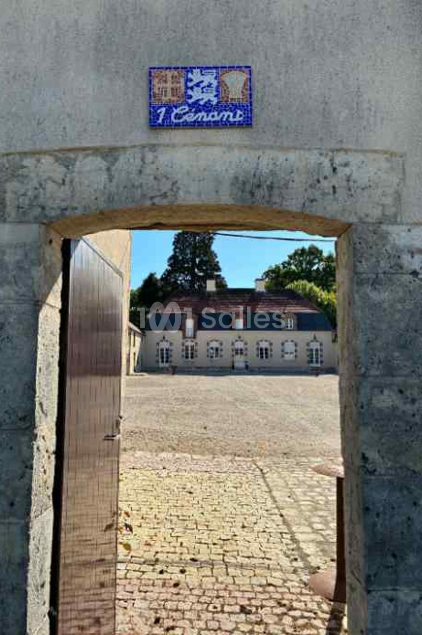 Vue à travers une arche en pierre ouverte sur une cour pavée et un bâtiment ancien avec des fenêtres blanches.