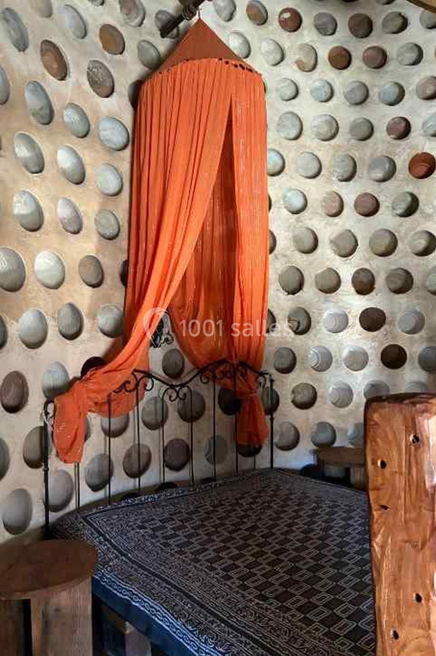 Lit en fer forgé avec moustiquaire orange suspendue, dans une pièce aux murs texturés avec des ouvertures circulaires.