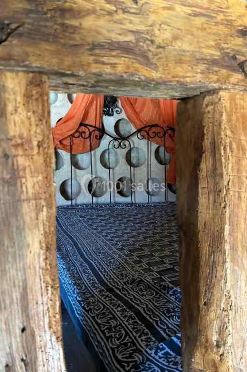 Vue à travers un cadre en bois brut d'un lit avec une couverture à motifs et un baldaquin orange en arrière-plan.