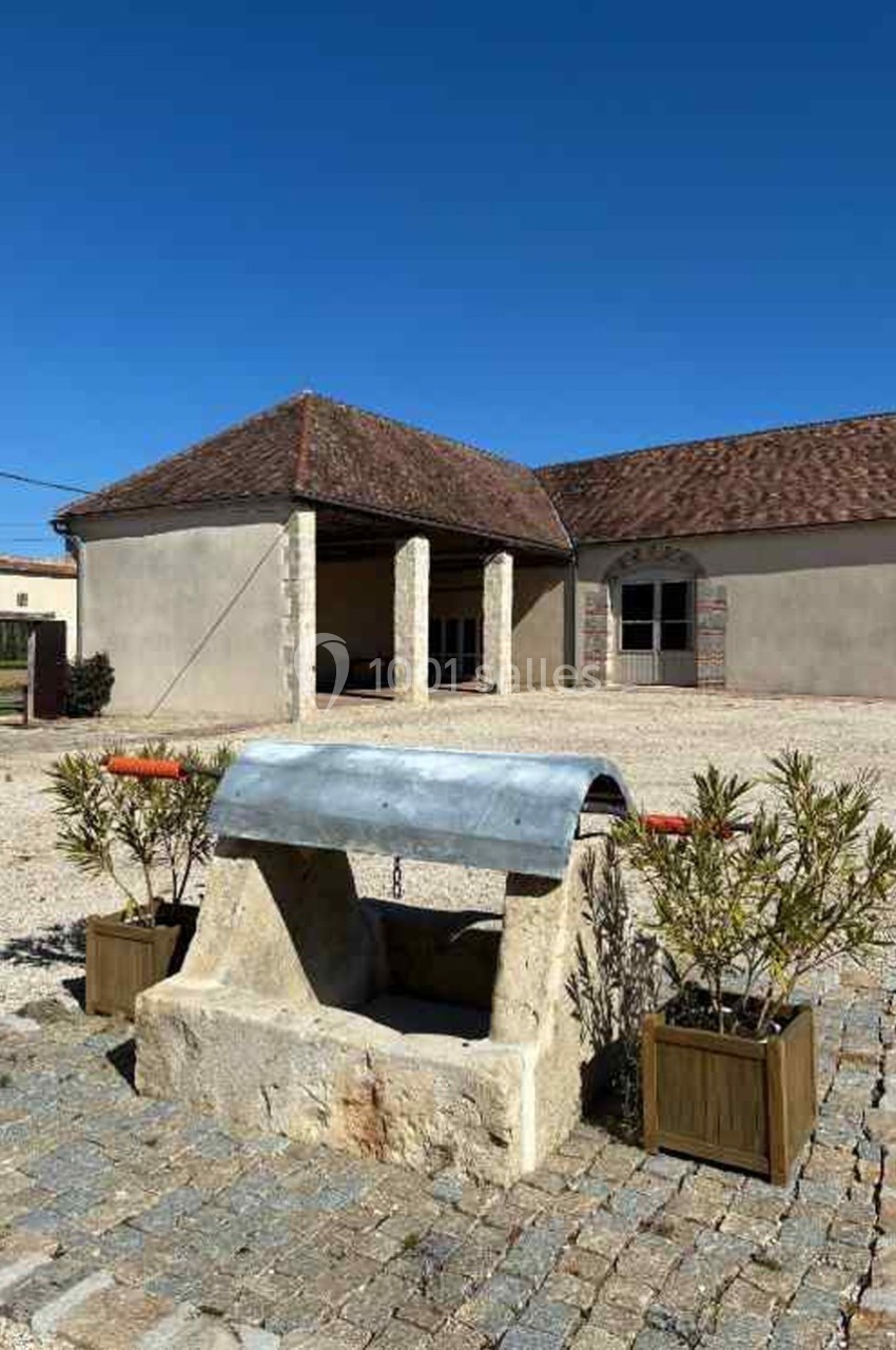 Cour pavée avec un puits en pierre au premier plan, entouré de bacs végétalisés, et bâtiments en arrière-plan.