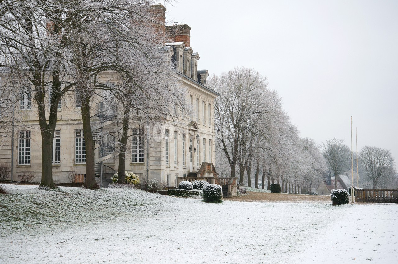 Façade d'un bâtiment ancien entouré d'arbres et d'un jardin recouvert de neige en hiver.