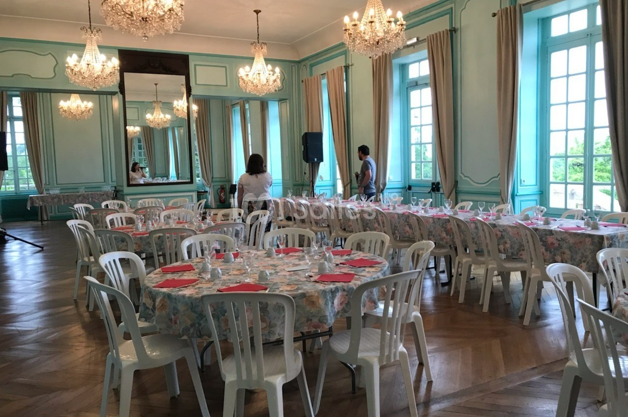 Salle de réception élégante avec tables rondes dressées, chaises blanches et lustres suspendus.