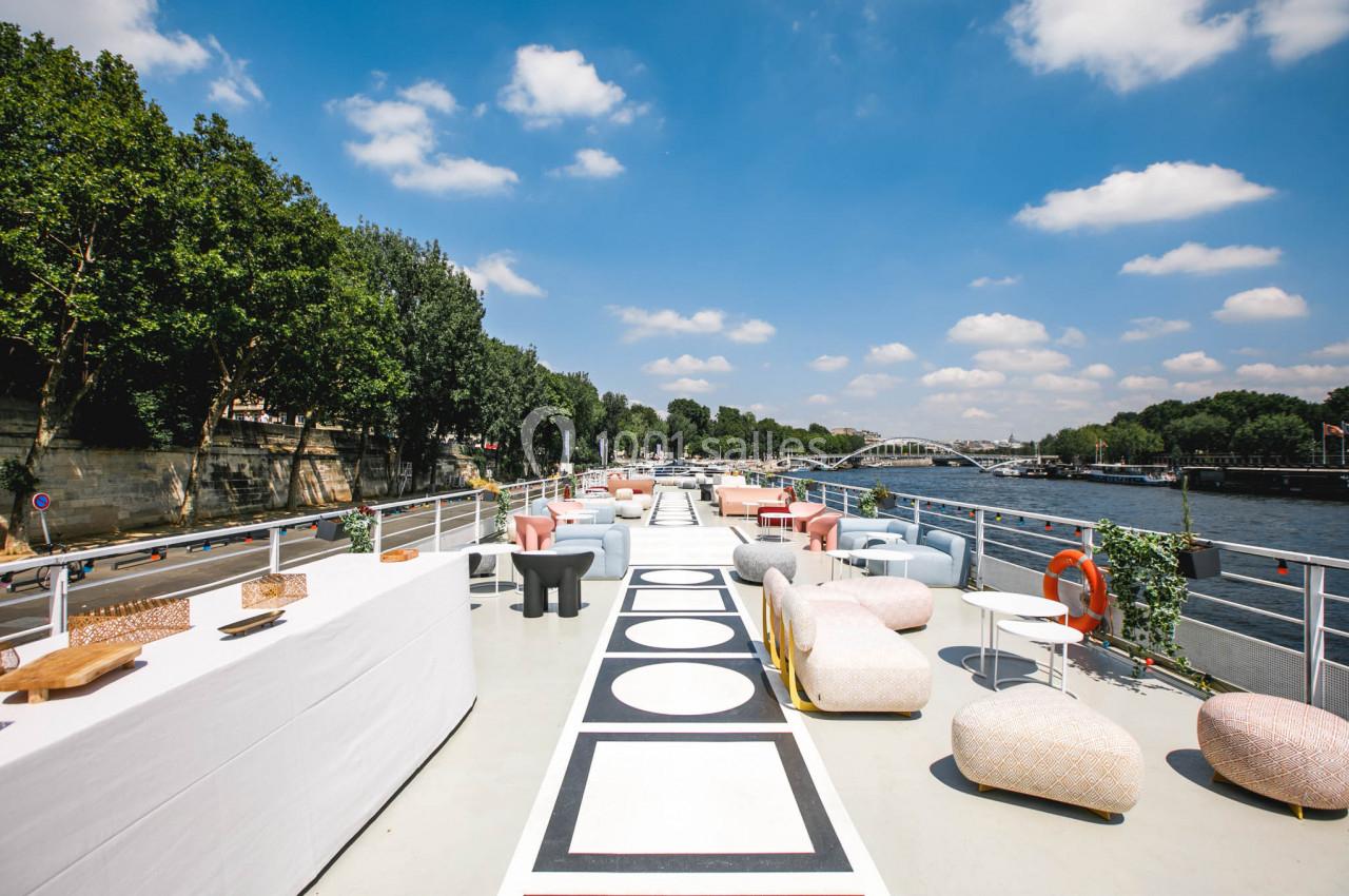Terrasse d'un bateau aménagée avec des tables et des fauteuils colorés, bordée par une rivière et des arbres.