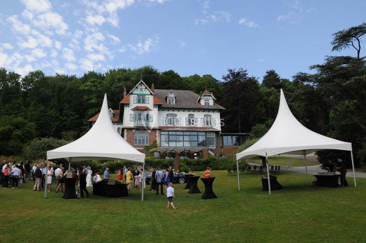 Des personnes rassemblées sous des tentes blanches sur une pelouse devant une grande maison entourée de verdure.