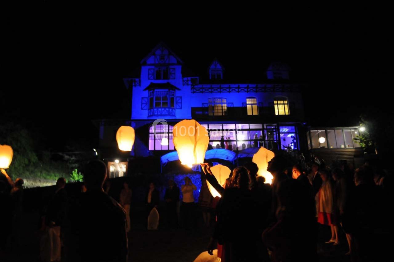 Des personnes lancent des lanternes lumineuses dans la nuit devant une grande maison éclairée en bleu.