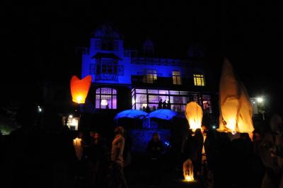 Miniature Location salle Cassel (Nord) - La Grande Maison Réception #34 Des lanternes lumineuses flottent dans la nuit près d'une grande maison éclairée en bleu, entourée de personnes.