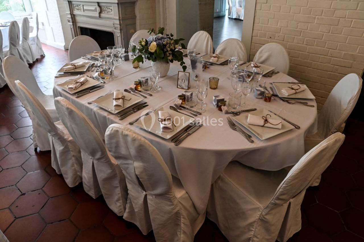 Table ronde dressée avec nappes blanches, vaisselle élégante, bouquets de fleurs et chaises couvertes.