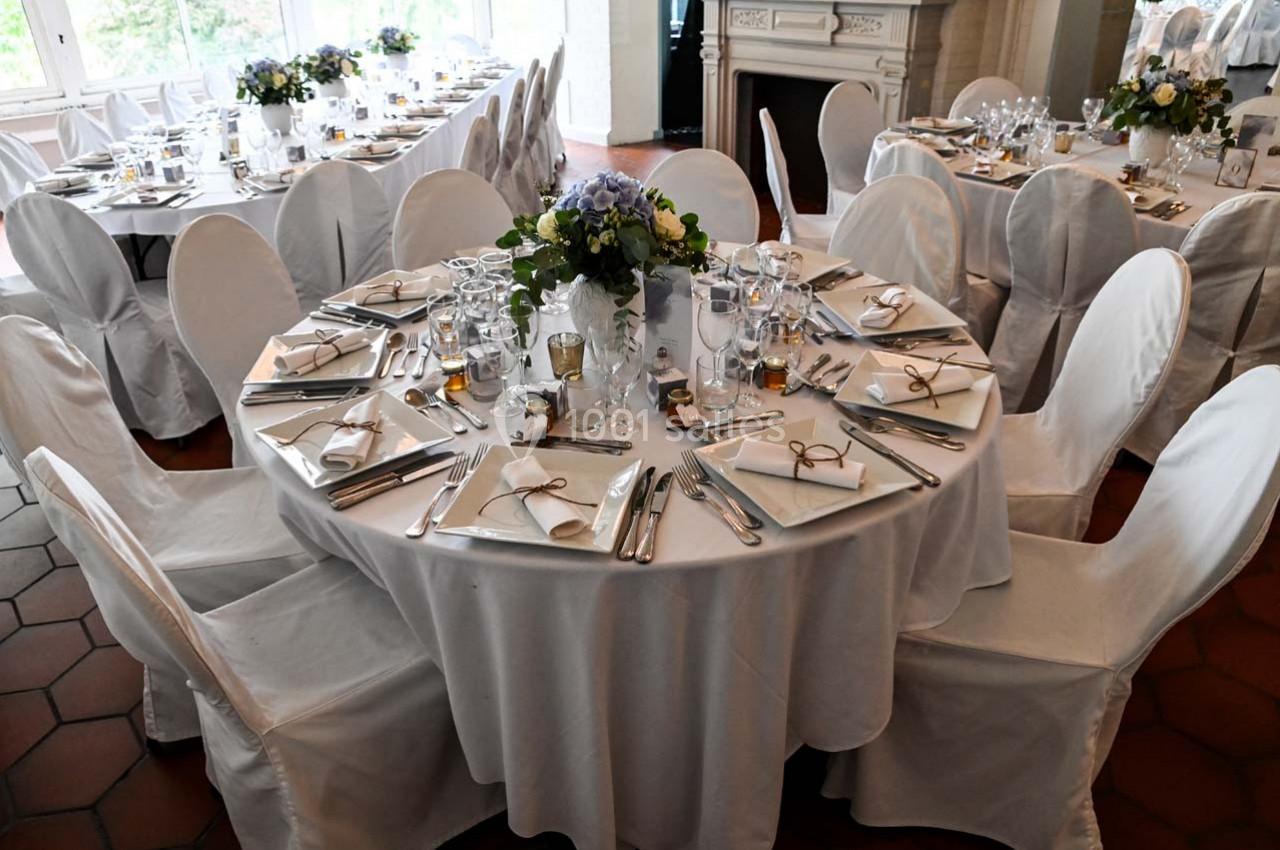 Table ronde élégamment dressée avec nappes blanches, couverts, verres et centre de table floral dans une salle lumineuse.
