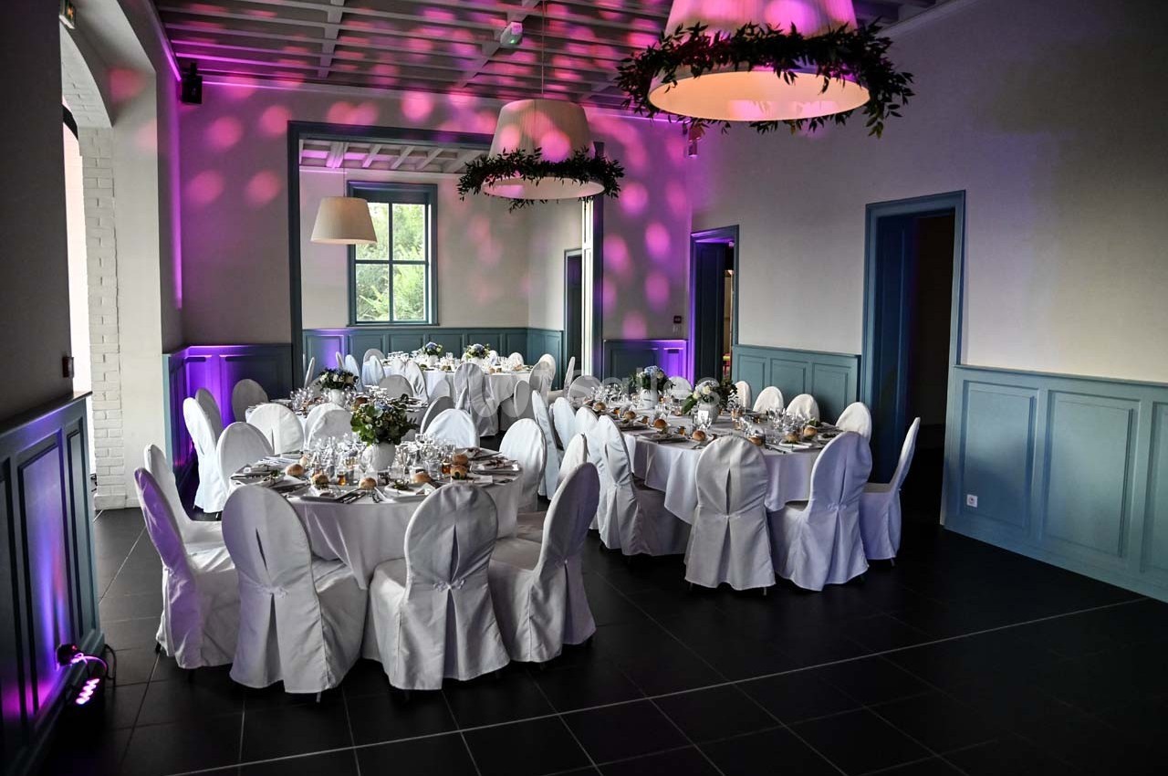 Salle de réception élégante avec tables rondes dressées, nappes blanches et éclairage d'ambiance violet.
