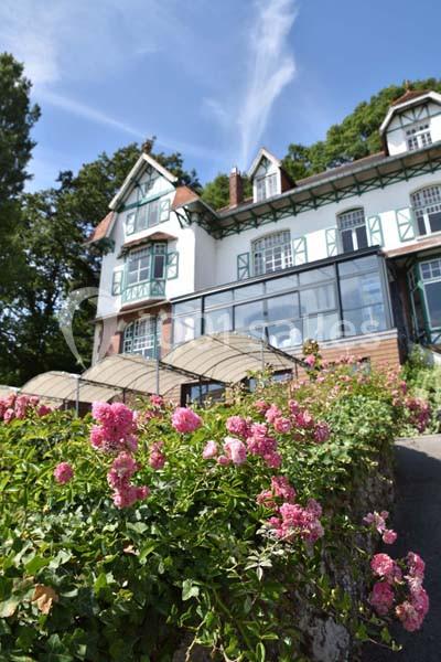 Grande maison blanche à colombages entourée de rosiers fleuris, sous un ciel bleu dégagé.