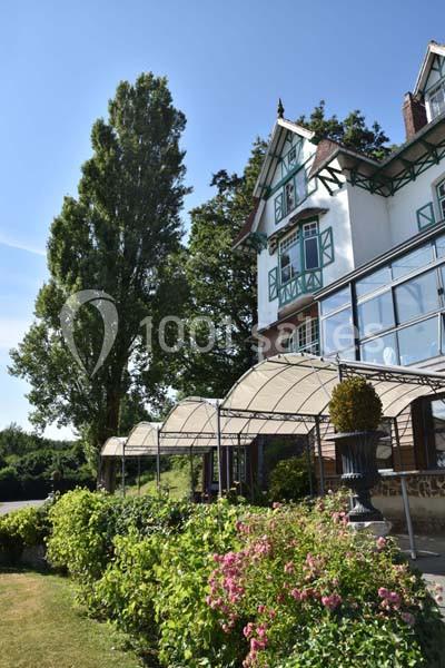 Façade d'une maison à colombages avec verrière, entourée de végétation et d'arbustes fleuris sous un ciel bleu.