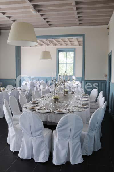 Salle de réception avec tables rondes dressées, nappes blanches, chandeliers et chaises couvertes de housses blanches.