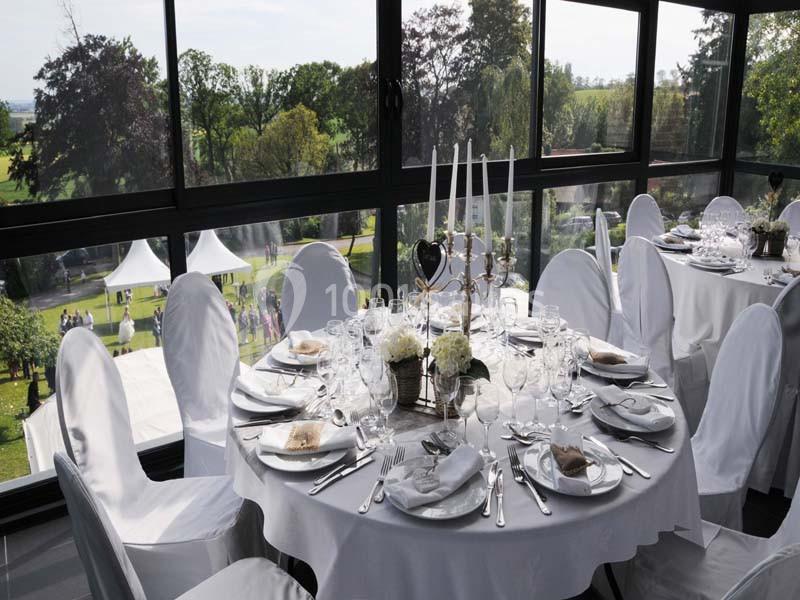 Salle lumineuse avec tables rondes élégamment dressées, nappes blanches, chandeliers et vue sur un jardin verdoyant.