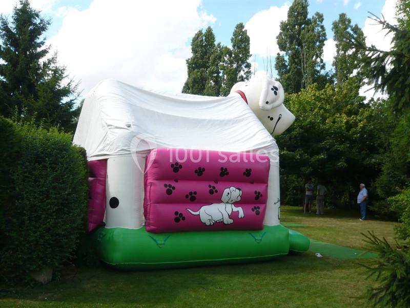 Structure gonflable en forme de chien blanc et rose installée dans un jardin entouré d'arbres.