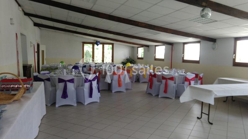 Salle de réception décorée avec des tables rondes, nappes blanches et chaises ornées de nœuds colorés.