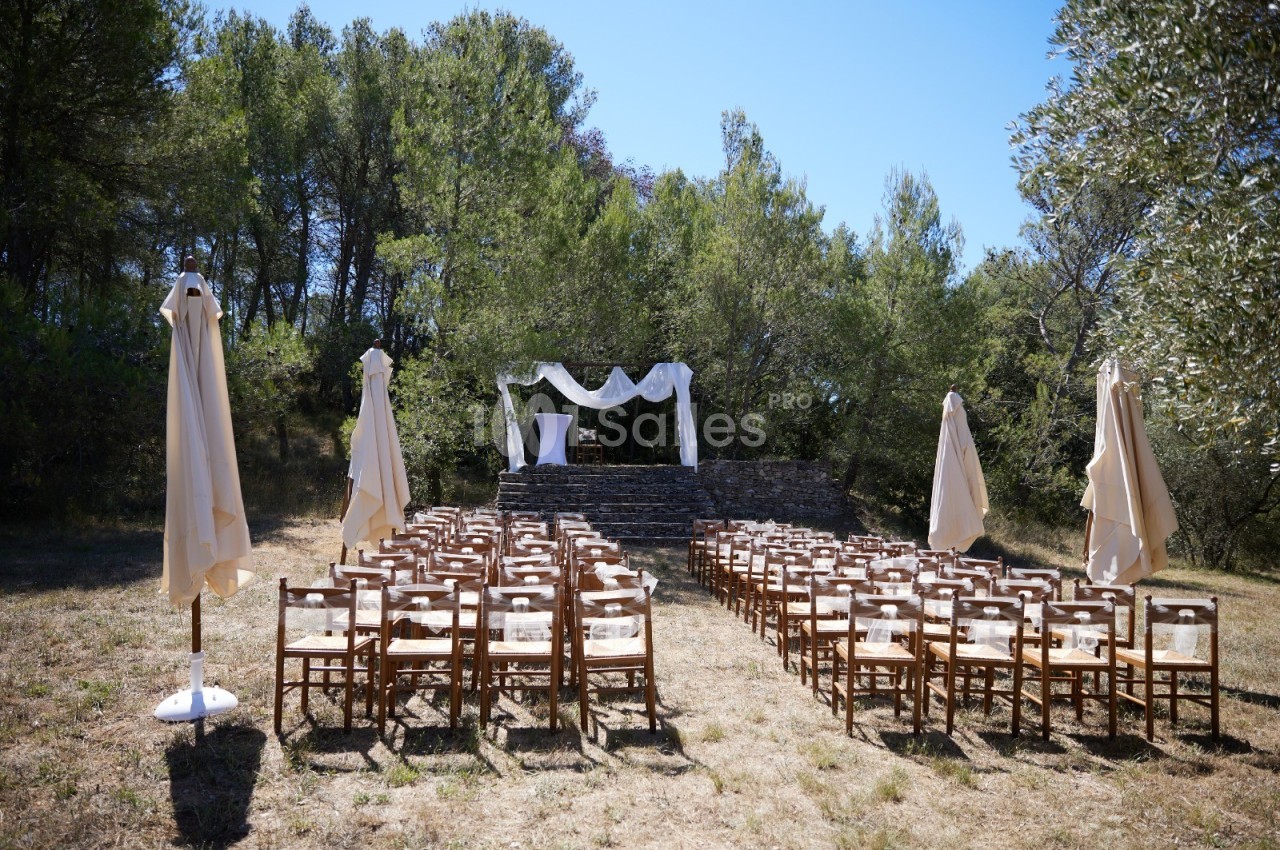 Chaises en bois disposées en rangées face à une arche décorée pour une cérémonie en extérieur, entourée de végétation.