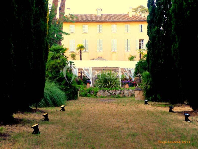 Location salle La Cadière-d'Azur (Var) - Château de Rochebelle - Var #3 Allée bordée d'arbres menant à une grande maison jaune avec une serre et un jardin verdoyant à l'arrière-plan.