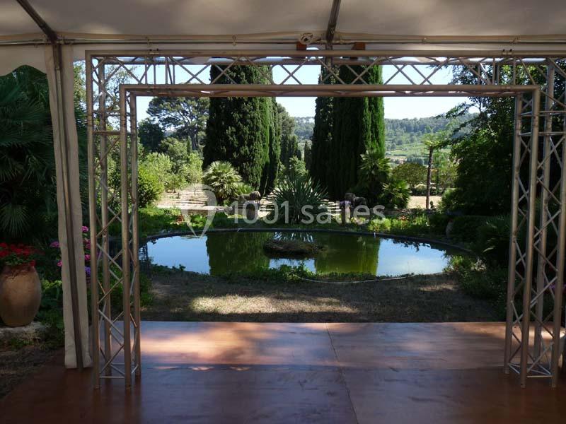 Location salle La Cadière-d'Azur (Var) - Château de Rochebelle - Var #16 Vue d'un jardin avec un bassin rond entouré de végétation, encadré par une structure métallique sous une tente.