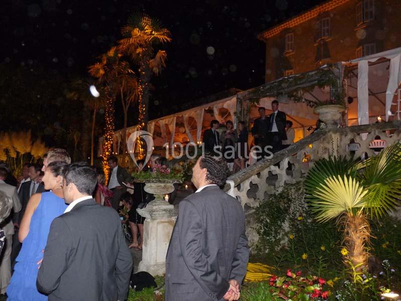 Location salle La Cadière-d'Azur (Var) - Château de Rochebelle - Var #17 Groupe de personnes en tenue de soirée discutant devant un bâtiment éclairé et entouré de végétation, de nuit.