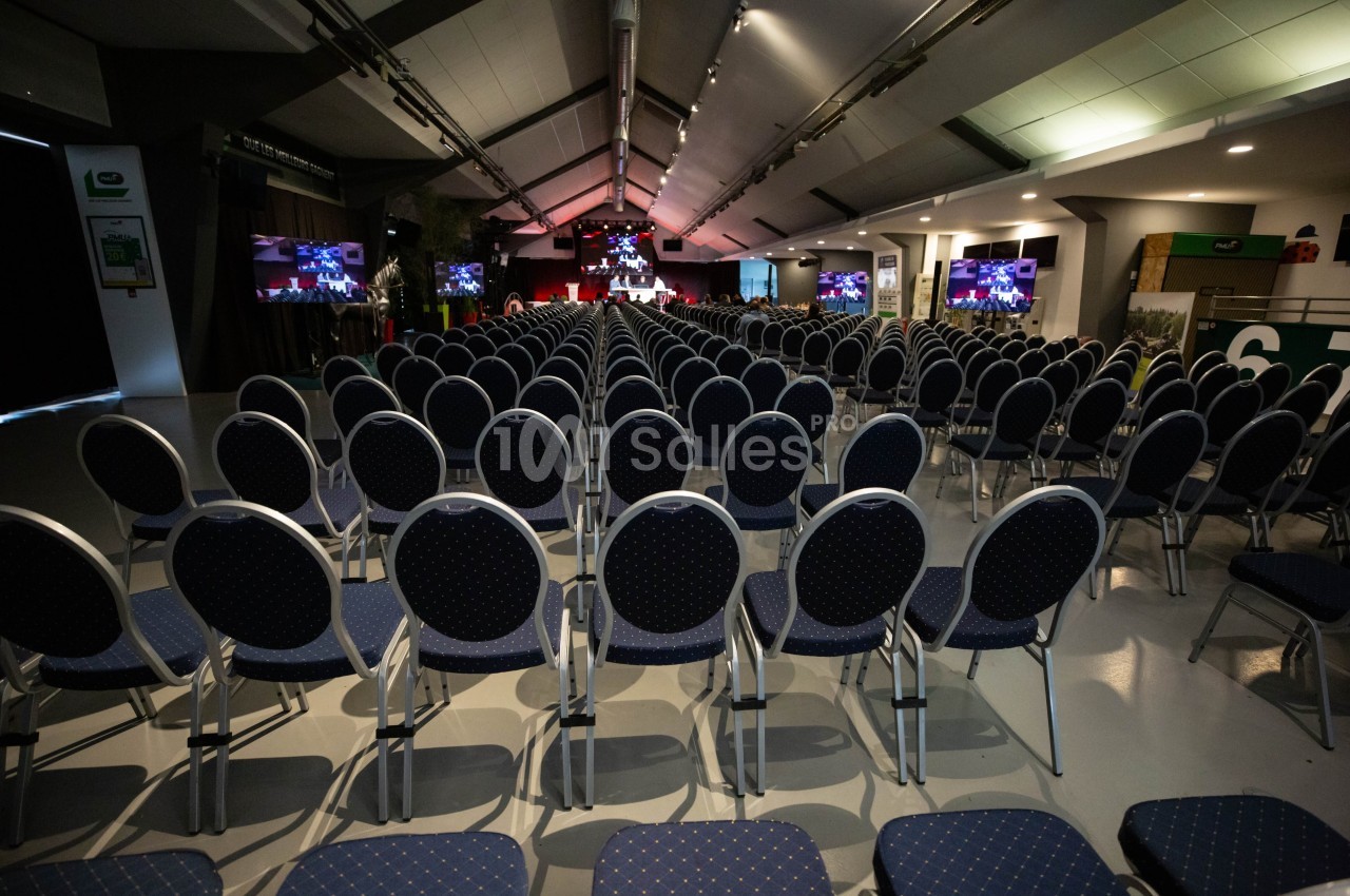 Salle de conférence avec des rangées de chaises vides, scène éclairée et écrans affichant des présentations.