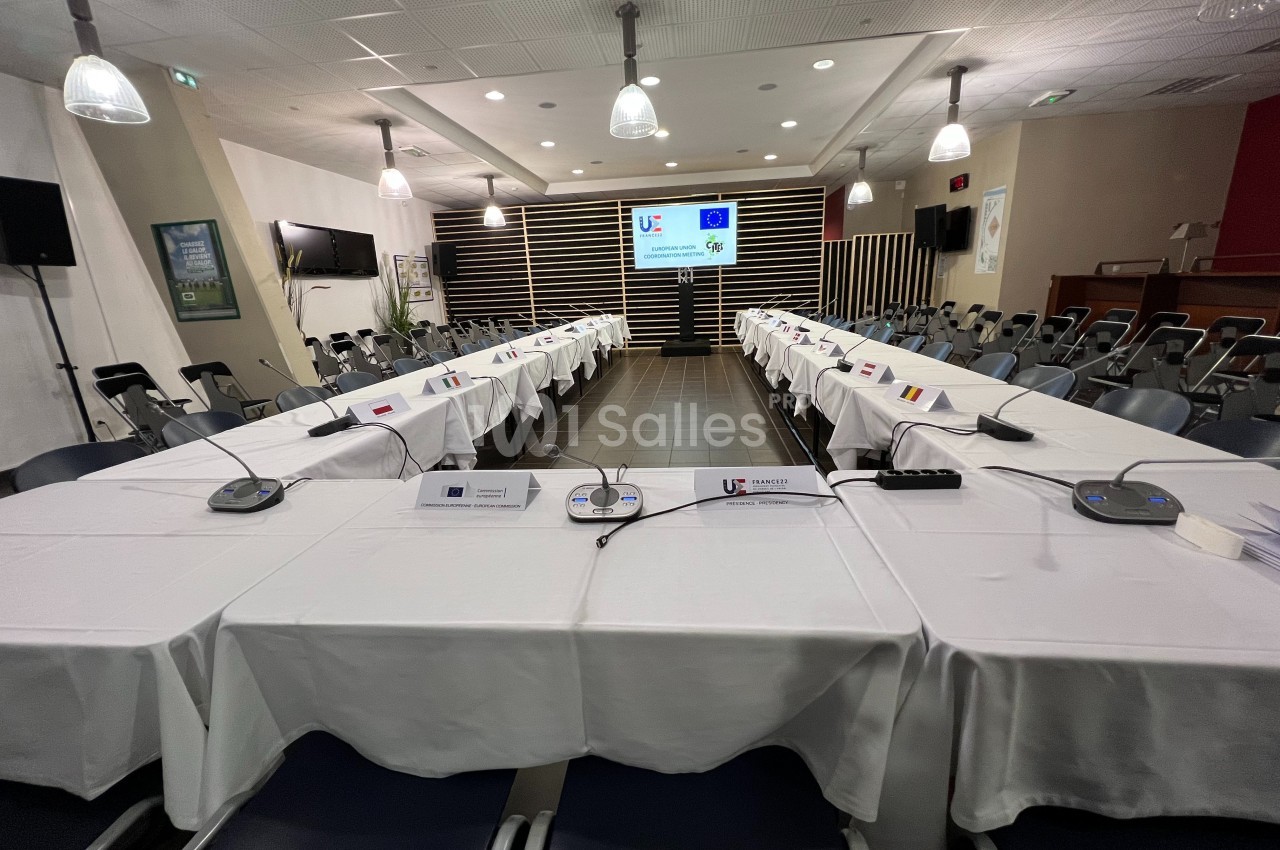 Salle de conférence avec tables disposées en U, microphones, écrans projetant des logos et chaises alignées.