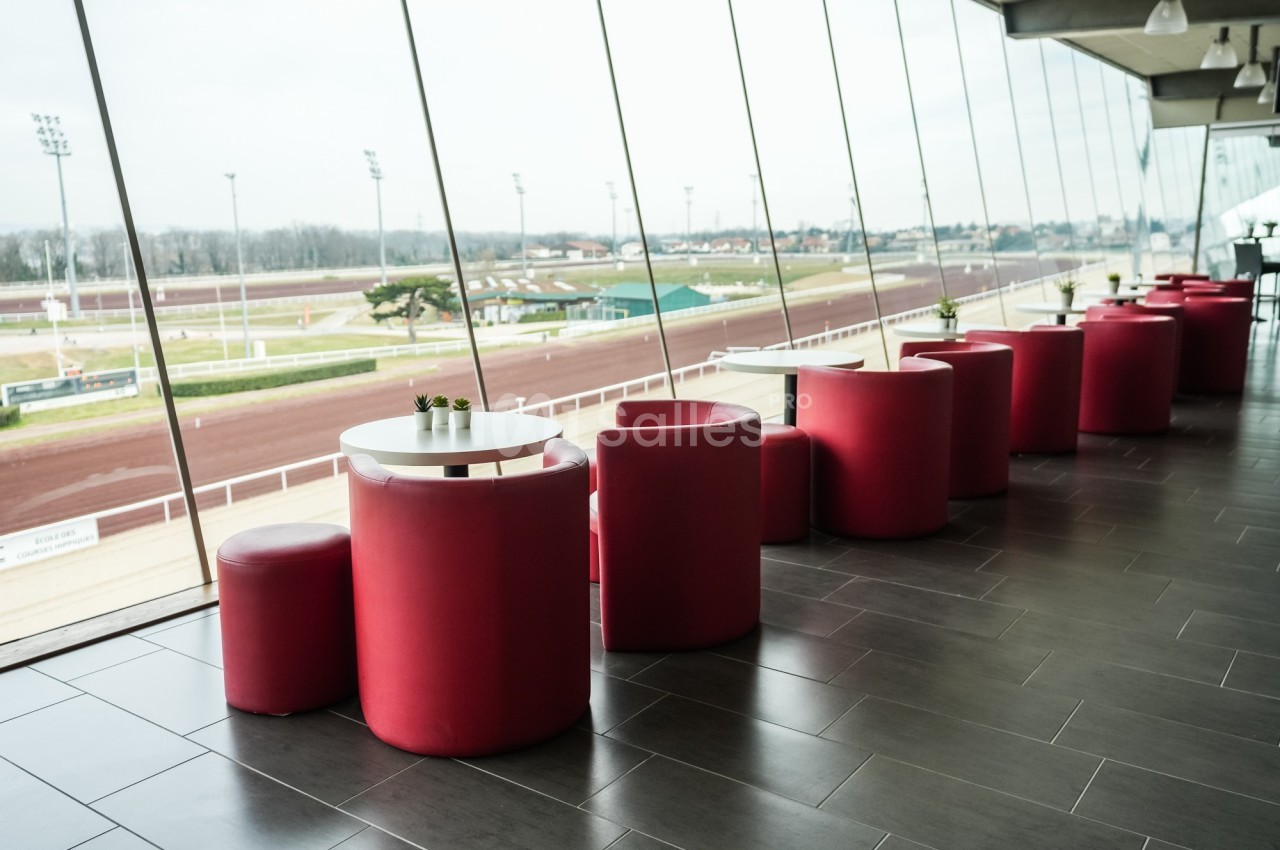 Salle avec des fauteuils rouges et des tables rondes, offrant une vue sur un hippodrome à travers de grandes baies vitrées.