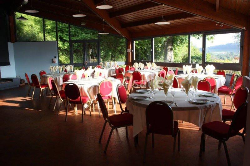 Salle lumineuse avec de grandes baies vitrées, tables rondes dressées avec nappes blanches et chaises rouges.