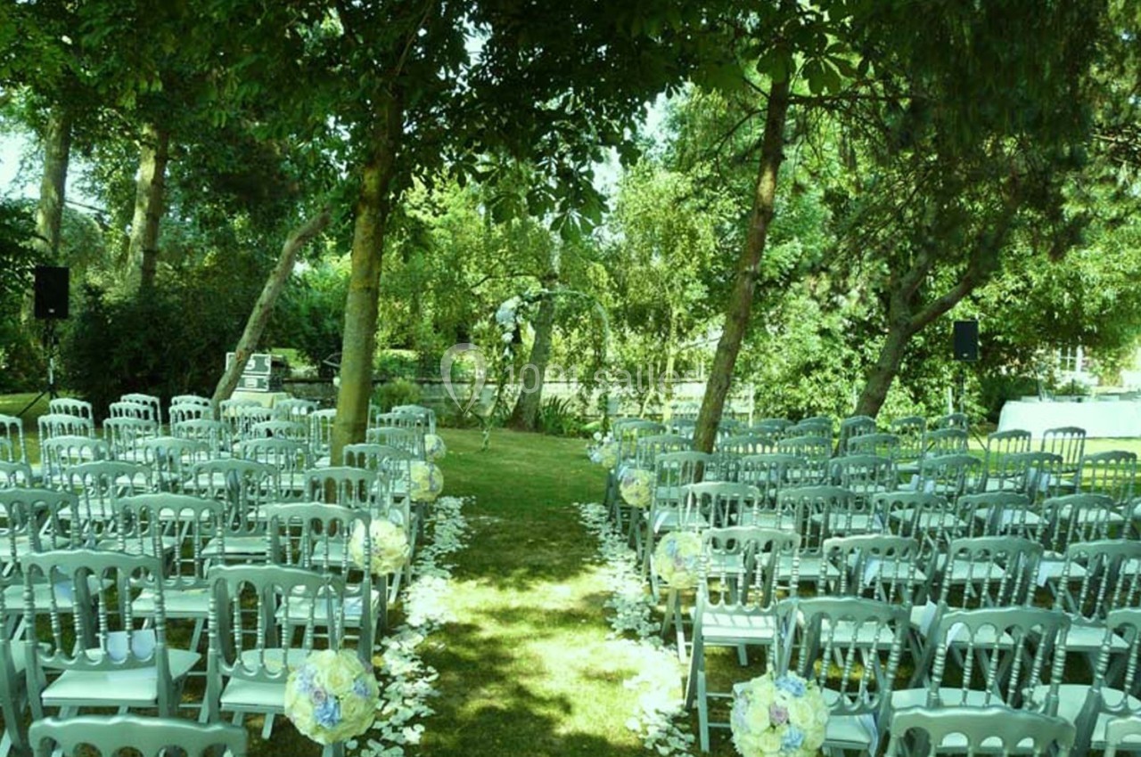 Chaises blanches alignées en extérieur sous des arbres, formant une allée décorée de fleurs pour une cérémonie.